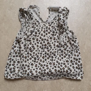 Topp - Säljer en söt ärmlös topp från H&M i storlek 68 med leopardmönster i svart och brunt på beige botten. Toppen har volangdetaljer vid axlarna och en knapp i nacken för enkel påklädning. Perfekt för dig som gillar djurmönster och vill sticka ut.