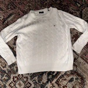 Vit kabelstickad tröja från GANT - Vit kabelstickad tröja från GANT med rund hals och diskret logga på bröstet. Tröjan har ribbade muddar vid ärmslut och nederkant, och är långärmad. Perfekt för dig som gillar klassisk och clean stil.
