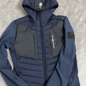 Mörkblå dunjacka Sail Racing strl S - Snygg mörkblå dunjacka från Sail Racing med huva och två bröstfickor med dragkedja. Jackan har quiltad framsida och rygg, samt detaljer i svart. Perfekt för kalla dagar med 800 FP dunfyllning. Logga på bröstet och ärmen.