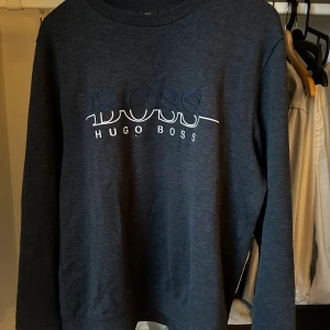 Mörkgrå sweatshirt från Hugo Boss - Snygg mörkgrå sweatshirt från Hugo Boss med stor BOSS-logga i blått och vitt på bröstet. Tröjan har rund hals och långa ärmar, perfekt för en chill och stilren look. Materialet är mjukt och bekvämt, passar dig som gillar klassisk streetwear.