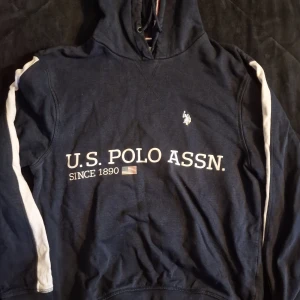 Mörkblå hoodie U.S. Polo Assn XS - Mörkblå hoodie från U.S. Polo Assn. i storlek XS med vit logga och text framtill. Tröjan har huva med snörning och vita ränder längs ärmarna. Materialet är mjuk bomull och polyester, perfekt för chill dagar. Klassisk design med broderad emblem på bröstet.