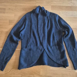 Mörkgrå ribbad kofta från H&M - Säljer en mörkgrå, öppen kofta från H&M Basic i storlek M. Koftan har snygga ribbade partier framtill och på ärmarna, samt en omlottliknande framkant. Perfekt för lager-på-lager och chill vardagsstil.