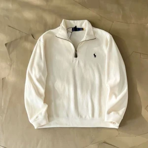 Vit half zip tröja från Ralph Lauren - Säljer en stilren vit långärmad tröja från Ralph Lauren med half zip och klassisk broderad logga på bröstet. Tröjan har hög krage och är tillverkad i mjuk bomull, perfekt för lager-på-lager under höst och vår.