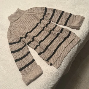 Beige och svart randig polotröja - Säljer en beige stickad polotröja med svarta ränder. Tröjan har grovstickad struktur, lång ärm och bred polokrage. Slits på båda sidorna. Använd 1 gång. 