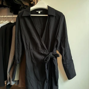 Svart omlottklänning med knyt - 🤍Svart omlottklänning med lång ärm, klassisk krage och knyt i midjan. Klänningen har en v-ringad front och är tillverkad i bomull/linne 🤍