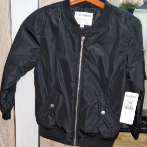 Svart bomberjacka från KappAhl - Svart bomberjacka från KappAhl med klassisk ribbad krage och muddar. Jackan har silverfärgad dragkedja, två fickor med tryckknappar och en extra ficka på ärmen. Materialet är glansig polyester och insidan är quiltad för extra komfort.