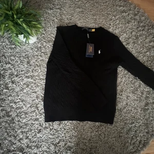 Svart kabelstickad tröja Polo Ralph Lauren - Svart kabelstickad tröja från Polo Ralph Lauren med klassisk rund hals och broderad logga på bröstet. Tröjan har lång ärm och ribbade muddar. Perfekt för dig som gillar stilrena och tidlösa plagg med premiumkänsla.