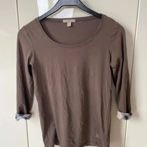 Brun långärmad topp från Burberry Brit - Snygg brun långärmad topp från Burberry Brit i mjuk bomull. Ärmarna har klassiskt rutigt tyg på insidan som syns när du viker upp dem. Rund halsringning och diskret Burberry-logga nertill. Passar även en small. 