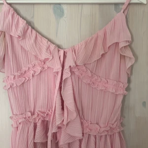 Rosa volangklänning från Amisu/new Yorker - Supersöt rosa klänning från Amisu med tunna axelband, volanger och plisserad mesh. Klänningen har omlottkänsla och romantiska detaljer som ger en drömmig vibe. Perfekt för dig som älskar pasteller och feminina plagg.