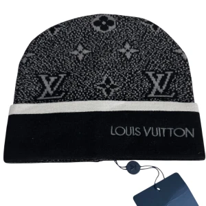Louis Vuitton svart och grå mössa - Exklusiv svart och grå mössa från Louis Vuitton med klassiskt LV-monogram och logotyp framtill. Mössan har en bred, ljusgrå kant och är tillverkad i ett mjukt, stickat material som håller dig varm och snygg. Perfekt accessoar för dig som vill sticka ut med lyxig streetstyle.