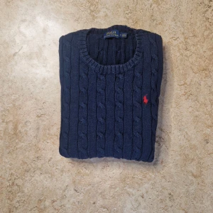 Marinblå stickad tröja Polo Ralph Lauren - Snygg marinblå stickad tröja från Polo Ralph Lauren med klassiskt kabelstickat mönster och röd broderad logga på bröstet. Rund halsringning och ribbade muddar vid ärmslut och nederkant. 