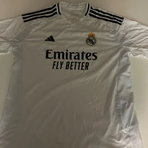 Real Madrid vit fotbollströja Adidas - Säljer en officiell Real Madrid fotbollströja från Adidas i storlek 2XL. Tröjan är vit med svarta detaljer, har korta ärmar och V-ringad krage. Broderat klubbmärke på bröstet och 'Emirates Fly Better' tryckt framtill. Materialet är lätt och andas, perfekt för match eller träning.