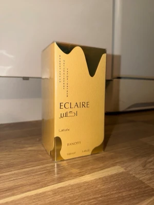Lattafa Eclaire Banoffi parfym - Eclaire Banoffi från Lattafa är en parfym i en lyxig guldfärgad förpackning med elegant design och droppformad detalj upptill. Flaskan rymmer 100 ml och har en modern, stilren look som passar dig som gillar exklusiva dofter.