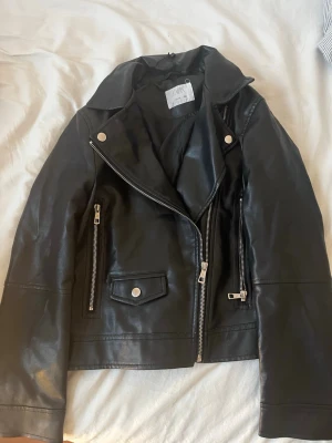 Svart biker skinnjacka från Zara - Snygg svart biker skinnjacka från Zara med silvriga dragkedjor och knappar. Klassisk modell med bred krage och två dragkedjefickor framtill. Perfekt för dig som vill ha en edgy och stilren look.