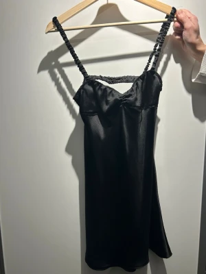 Svart satin klänning med axelband - Snygg svart klänning i satin med glansig finish och unika, rynkade axelband. Klänningen har en figurnära passform upptill och lätt utställd nederdel. Perfekt för dig som gillar stilrena och trendiga plagg med en twist.