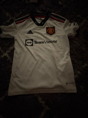 Manchester United bortatröja Adidas - Snygg vit Manchester United bortatröja från Adidas med svarta och röda detaljer. Tröjan har klubbmärke på bröstet, Adidas-logga och TeamViewer sponsortryck. Tillverkad i lätt och ventilerande material, perfekt för fotboll eller träning.