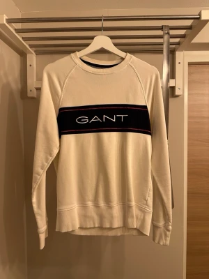 Vit sweatshirt från GANT - Snygg vit sweatshirt från GANT med klassisk rund halsringning och ett brett mörkblått band över bröstet där GANT-loggan är tryckt i vitt. Tröjan har långa ärmar, ribbade muddar och är tillverkad i mjukt bomullsmaterial. Perfekt för en avslappnad och stilren look.