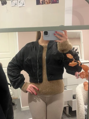 Svart croppad bomberjacka - Säljer en svart croppad bomberjacka med dragkedja framtill. Jackan har en klassisk bomberform med ribbade muddar och lång ärm. Perfekt för dig som vill ha en stilren och trendig jacka till vardagslooken.