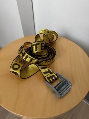 Gult Off-White industribälte - Säljer ett gult industribälte från Off-White med svart text och metallspänne. Bältet har den ikoniska loggan och texten längs hela bandet. Perfekt för att ge outfiten en streetwear-vibe och sticker verkligen ut med sin färg och design. 