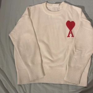 Vit stickad tröja från Ami Paris - Vit stickad tröja från Ami Paris. Röd detalj. Liten färgfläck på baksidan av högra armhålan, därav priset! Hör gärna av er vid frågor eller bud!