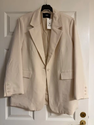 Beige kavaj från Bik Bok - Stilren beige kavaj från Bik Bok med klassisk krage, två framfickor med lock och knäppning framtill. Kavajen har långa ärmar med dekorativa knappar vid ärmsluten och en rak passform som ger en snygg siluett.