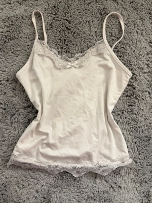 Vitt pyjamaslinne med spets och en rosett.  - Ett fint pyjamaslinne med spets upptill och nedtill. Även en rosett (om du inte gillar den så klipp bort den) där framme. Väldigt mjukt material och säljer för att det aldrig används. Endast gjort någon enstaka gång. Vet inte storlek eller märke🤍