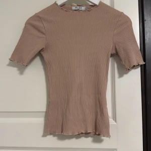 Beige ribbad topp från NA-KD - Supersnygg beige ribbad topp från NA-KD med korta ärmar och vågig kant vid hals, ärmslut och nederkant. Figurnära passform och stretchigt material som sitter perfekt. Perfekt att styla med jeans eller kjol för en trendig look.