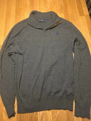 Grå stickad tröja från Polo Ralph Lauren - Mörkgrå stickad tröja från Polo Ralph Lauren med halv dragkedja och hög krage. Tröjan har en blå broderad logga på bröstet och ribbade muddar vid ärmslut och nederkant. Perfekt för lager på lager och stilren look. Det är en lite defekt på ärmen men det syns knappt