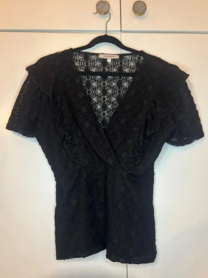Svart kortärmad spetsblus med volang - Svart mönstrad blus med volanger och v ringning där framme. Passar för utgång eller om man vill vara extra fin! Endast använd 2 gånger