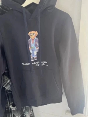 Svart hoodie Polo Bear Ralph Lauren - Svart hoodie från Ralph Lauren med Polo Bear-tryck framtill. Klassisk huva med dragsko och känguruficka. Snygg detalj med björnen i färgglada kläder och texten 'Polo Bear by Ralph Lauren' under motivet. Perfekt för en trendig och avslappnad stil.