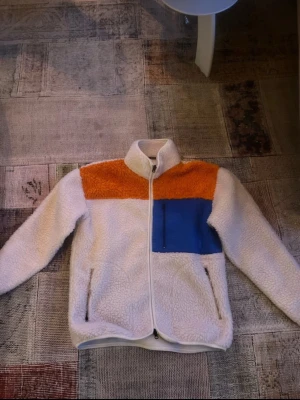 Teddyjacka från Aveny med färgblock - Säljer en mysig teddyjacka från Aveny med vit bas, orange axelparti och blå bröstficka. Jackan har hel dragkedja framtill, två sidofickor och hög krage. Perfekt för dig som gillar färg och vill ha en varm jacka till hösten eller vintern. Nypris: 1899kr