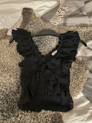 Svart spetsvolang topp från Zara - Supersnygg svart topp från Zara med volanger och spetsdetaljer. Djup v-ringning och breda axelband ger en trendig look. Perfekt att styla med jeans eller kjol för en cool outfit.