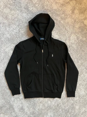 Svart kofta - Snygg svart hoodie från Polo med dragkedja framtill, två fickor och klassisk huva med dragsko. Ikoniska Polo-loggan broderad på bröstet. Tillverkad i mjukt bomullsmaterial som är skönt att bära. Perfekt för en avslappnad och stilren look.