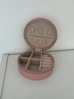 Rosa oval smyckesväska med fack - Söt oval smyckesväska i ljusrosa syntetmaterial med dragkedja. Insidan har flera fack och ringhållare, samt en liten ficka med resår. Perfekt för att organisera smycken när du är på språng.