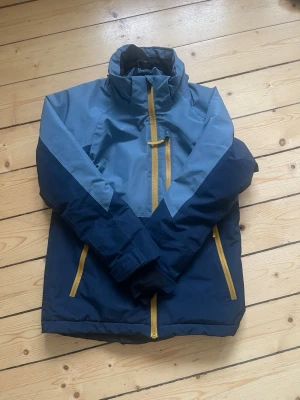 Blå vindjacka från Everest - Snygg tvåfärgad blå vinterjacka från Everest med gula dragkedjor. Storlek 146. Jackan har hög krage, långa ärmar och praktiska fickor framtill. Perfekt för blåsiga dagar och outdoor-aktiviteter. 