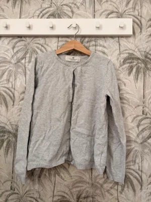 Ljusgrå cardigan från H&M - En enkel och stilren ljusgrå cardigan från H&M med rund halsringning och knäppning framtill. Tillverkad i mjukt material och har långa ärmar samt ribbade muddar vid ärmslut och nederkant. Perfekt att slänga över en t-shirt för en avslappnad look.