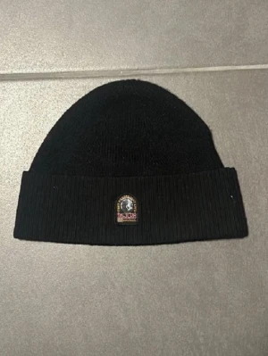  mössa Parajumpers - Snygg svart ribbstickad mössa från Parajumpers med uppvikt kant och klassisk PJS-logga framtill. Perfekt för kalla dagar och enkel att matcha med olika outfits. Tillverkad i mjukt material som sitter skönt på huvudet.