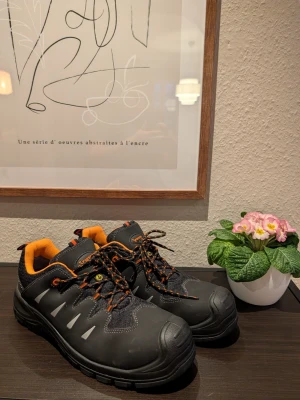 Svarta sneakers från Gesto med orange detaljer - Arbetsskor svarta sneakers med stålhätta från Gesto med orange detaljer och snörning. Skorna har en robust design med rund tå, platt sula och inslag av syntetmaterial. Insidan är fodrad i orange och skosnörena är svart-orange mönstrade för extra stil.