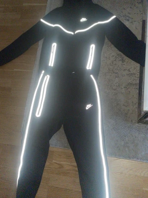 Svart Nike loungewear-set med reflex - Svart loungewear-set från Nike med vita reflexdetaljer längs dragkedjor och sömmar. Setet består av en hoodie med dragkedja och matchande byxor, båda med Nike-logga. Perfekt för chill dagar hemma eller när du vill sticka ut med coola reflexer.