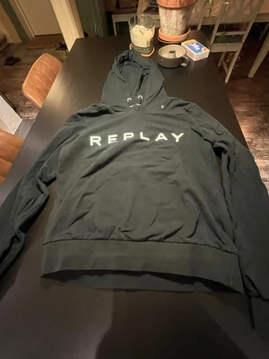 Svart hoodie från Replay - Snygg svart hoodie från Replay med stor vit logga över bröstet. Klassisk modell med huva och dragsnören, samt ribbade muddar vid ärmslut och nederkant. Tillverkad i mjukt bomullsmaterial som är skönt att ha på sig.