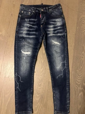 Mörkblå Dsquared2  - Säljer ett par mörkblå skinny jeans från Dsquared2 med slitningar, tryckta detaljer och coola tvättade effekter. Jeansen har klassisk femficksmodell, låg midja och smal passform. Perfekta för dig som gillar streetwear och vill sticka ut.