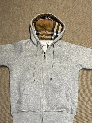 Grå zip hoodie från Burberry - Snygg grå zip hoodie från Burberry med klassiskt rutigt foder i huvan och broderad logga på bröstet. Tillverkad i mjuk bomullsblandning, har dragsko i huvan och två fickor framtill. Perfekt för en avslappnad och trendig look.