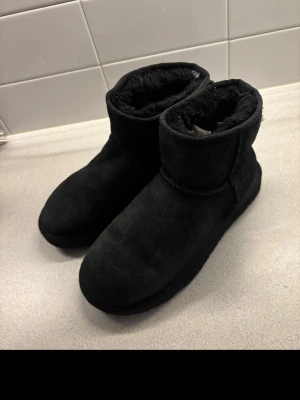 Svarta UGG  - Svarta  klassiska UGG i mjuk mocka med rund tå och fluffigt foder på insidan. Klassisk och enkel design med låg skaft och platt sula, perfekta för kyliga dagar. UGG-logga bak på hälen.