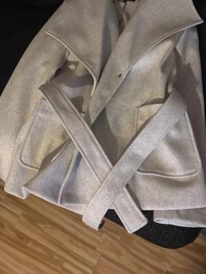 Beige  kappa med bälte - Stilren beige kappa med bred krage och stora fickor framtill. Kappan har ett matchande bälte i midjan för snygg passform och är perfekt för kyligare dagar. Enkel och modern design som passar till många olika outfits. Aldrig använd