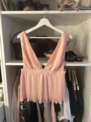 Plisserad ljusrosa omlottopp - Säljer en plisserad ljusrosa topp med djup v-ringning både fram och bak. Toppen har breda axelband, omlottdesign med knyt i ryggen och är ärmlös. Perfekt för dig som gillar en feminin och trendig stil. Det är ingen fläck på bild tre utan bar tyget som ser ut så, inget som syns när man bär toppen💓💓💓storlek m men sitter bra på mig som bär en s