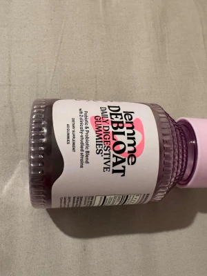 Lemme Debloat Digestive Gummies - Burk med Lemme Debloat Daily Digestive Gummies, ett kosttillskott med prebiotika och probiotika. Burken är genomskinlig med lila lock och etikett i vitt, svart och rosa. Innehåller 60 gummies för dagligt intag.