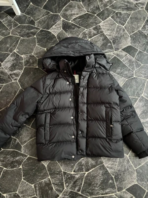 Svart Moncler jacka - Snygg svart dunjacka från Moncler med stor huva och quiltad design. Jackan har dragkedja och knappar framtill samt två praktiska fickor med dragkedja. Perfekt för kalla dagar och ger en riktigt clean look.