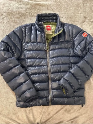 Mörkblå dunjacka från Colmar - Snygg mörkblå dunjacka från Colmar med quiltad design och dragkedja framtill. Jackan har hög krage, långärmad passform och diskret logga på ärmen. Perfekt för kalla dagar och enkel att matcha med olika outfits.