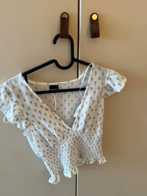 Blommig omlottopp från Gina Tricot - Supersöt vit omlottopp från Gina Tricot med små blå blommor. Toppen har korta ärmar, v-ringning och smockad midja som ger en snygg passform. Perfekt för dig som gillar romantiska detaljer och en feminin look.