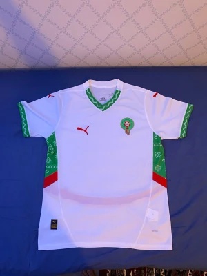 Marocko fotbollströja Puma vit/grön - Säljer en officiell Marocko fotbollströja från Puma i vitt med gröna och röda detaljer. Tröjan har korta ärmar, grönt mönster längs sidor och ärmslut samt Marockos landslagsemblem på bröstet. Perfekt för fotbollsfans!